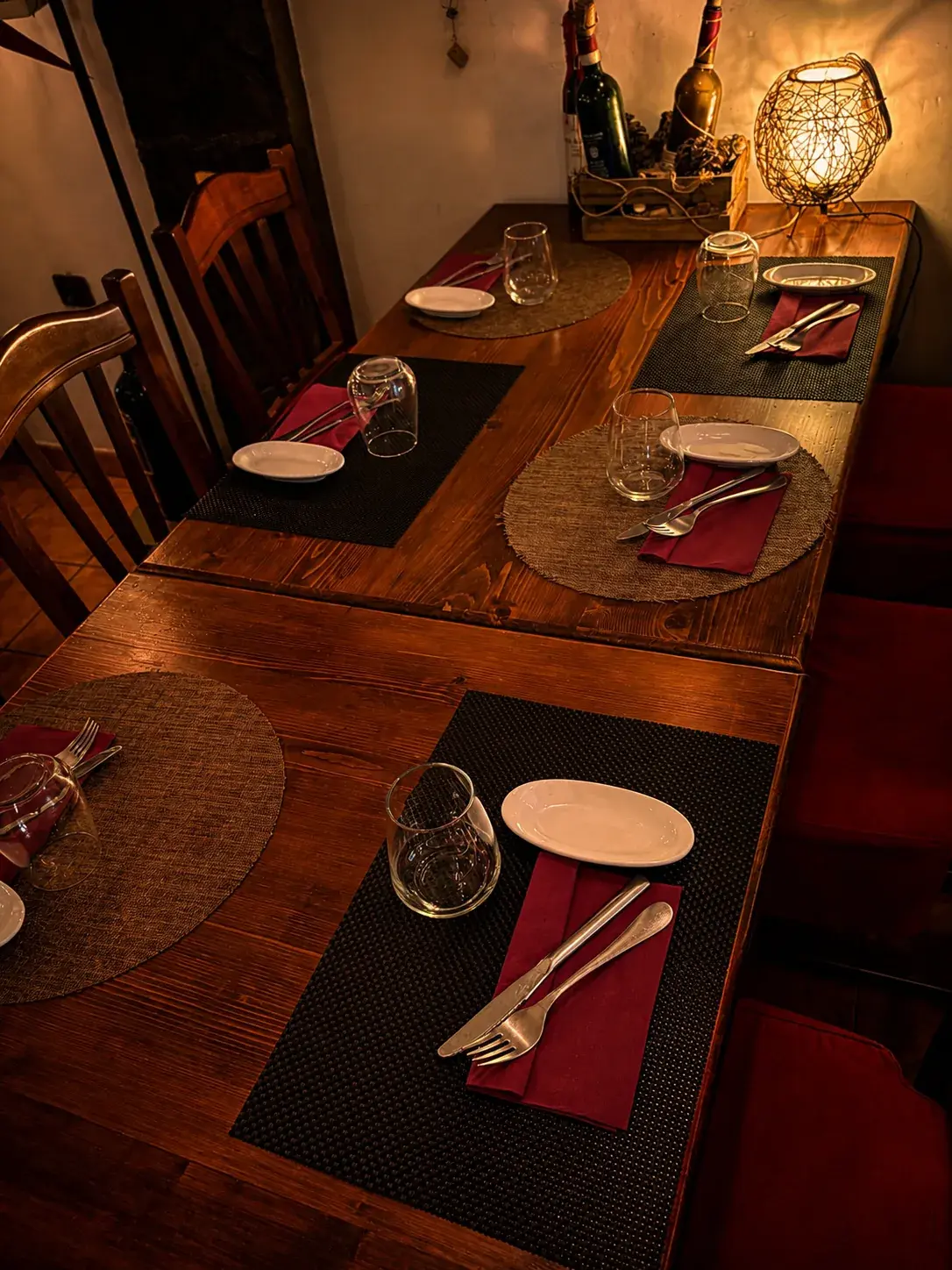 Wooden tables laid for dinner with woven and dark place mats, burgundy napkins and glasses, lit by a lantern set among wine bottles.